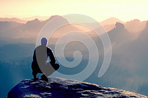 Young man in sportswear is sitting on cliff& x27;s edge and looking to misty valley bellow