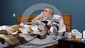 A young man sprays medicines in his nose. The guy is sick lying on the bed with a scarf around his neck.