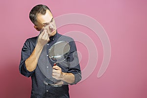 Young man squeezing pimple looking into mirror. Isolated on pink background. Acne. Skin care.