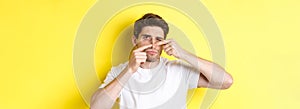 Young man squeezing pimple on nose, standing over yellow background. Concept of skin care and acne