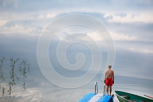 Young man standing alone on the jetty in the lake. Sunrise over the lake in the village