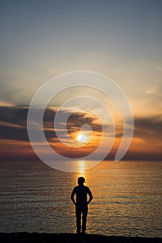 Young man standing at the beach in front of amazing sea view at sunset or sunrise and thinking about his future. Rear