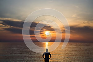 Young man standing at the beach in front of amazing sea view at sunset or sunrise and thinking about his future. Rear view