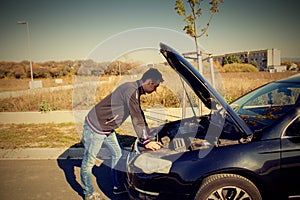 A young man is standing by a broken car and looks under the car hood.