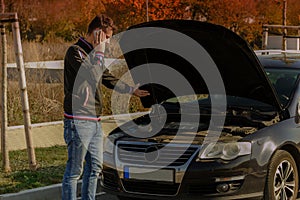 A young man is standing by a broken car and looks under the car hood.