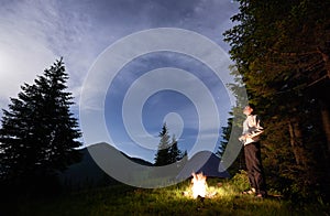 Young man is standing by a burning fire under evening sky with manifesting stars on the background of camping