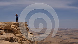 Young man standing on cliff edge and taking pictures of the desert on his phone