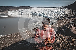 A young man standing in front of Eqip Sermia glacier called Eqi Glacier. Tourist holding an old analogue camera. Wall of
