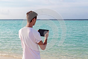 Young man standing in front of the sea and using his tablet during sunset