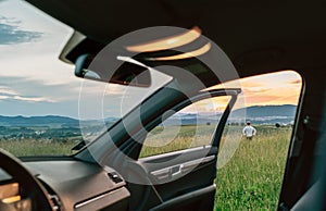 Young man standing in high grass and enjoying the sunset sky colors through the open car dor view. Traveling by car concept image