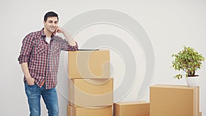 Young man standing in his new apartments, leaning against the pile of carton boxes, smiling, pointing at the copyspace