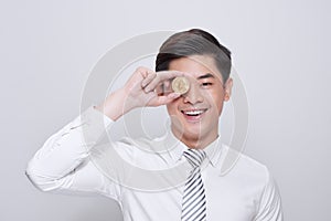 Young man standing isolated on white background holding cryptocurrency coin covering eye looking aside smiling playful
