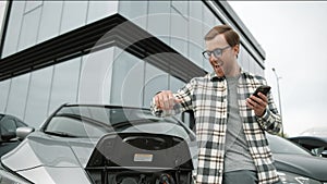 Young man standing near an electric car that is charging and making time adjustments on a smartphone