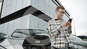 Young man standing near an electric car that is charging and making time adjustments on a smartphone