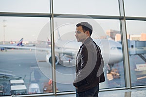 Young man is standing near window at the airport and watching plane before departure. Focus on his back