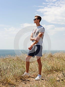 Young man standing and posing on top of cliff in summer mountains at sunset and enjoying view of nature.