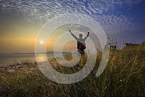 Young man standing and rising hand as victory on grass hill looking to sun above sea horizontal with dramatic colorful sky