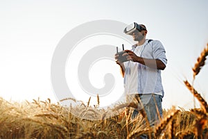 Young man standing in a wheat field at sunset in virtual reality glasses