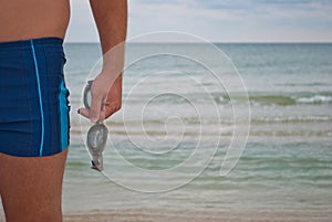 Young man stands against the sea shore beach sand waves water horizon holds in his hand sunglasses for swimming