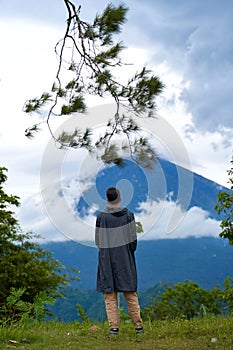 A young man stands with his back in a black raincoat enjoying the view of the popular sacred mountain Volcano Agung, shrouded in