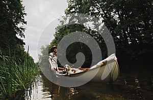 Young man stands in shallow river next to his canoe with a dog