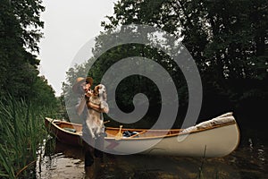 Young man stands in shallow river next to his canoe with a dog. Adventurous active rest concept