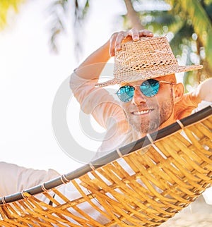 Young man in straw hat and blue sunglasses  lying swinging in cozy hammock sincerely smiling meeting morning sunrise sunlight on