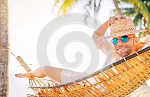 Young man in straw hat and blue sunglasses  lying swinging in cozy hammock sincerely smiling meeting morning sunrise sunlight on photo