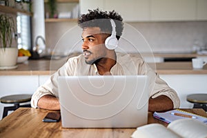 Young man student with laptop and headphones indoors at home, studying.