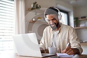 Young man student with laptop and headphones indoors at home, studying.