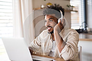 Young man student with laptop and headphones indoors at home, studying.