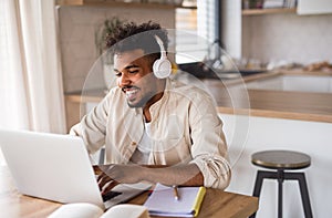 Young man student with laptop and headphones indoors at home, studying.