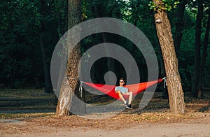 Young man in stylish casual clothes and sunglasses sitting in a hammock on a background of the park and admiring the sunset. Relax