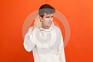 Young man with stylish hairdo in casual sweatshirt listening for, holding hand near his ear, interesting with gossips and secrets