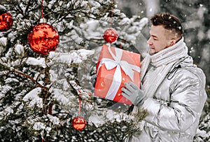 Young man in stylish silver jacket standing near decorated Christmas tree and holding holiday present box outdoors