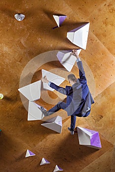 Young man in suit climbing difficult route on artificial wall in bouldering gym. Career challenges concept