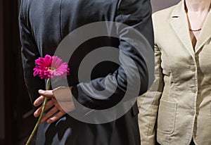 A young man in a suit holds a gerbera flower behind his back, a surprise for a woman, March 8