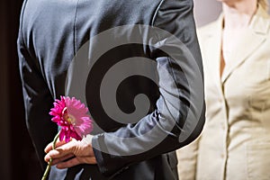 A young man in a suit holds a gerbera flower behind his back, a surprise for a woman, March 8