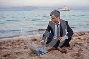 young man in suit with laptop sitting on the beach working on the seaside