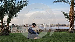 Young man in suit with laptop working on green grass near a resort on the seaside