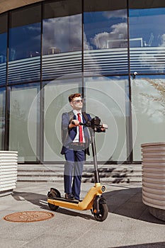 Young man in suit rides on electric scooter in business district at modern corporate building.