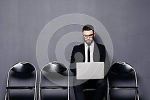 Young man in suit sitting on chair with laptop and waiting for job interview against gray background