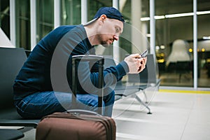 A young man with a suitcase sits in the airport waiting room and uses a mobile phone. Night flight, transfer, waiting at
