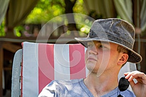A young man in the summer in a hat on a lounge chair on a clear sunny day, against the background of a summer gazebo, is resting