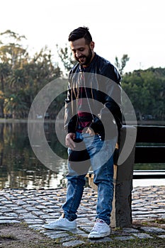 happy Latin man in a leather jacket and blue jean pants standing by a lake