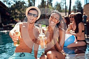 Young man is sunglasses and cheerful woman cheering with beer bottles in swimming pool.