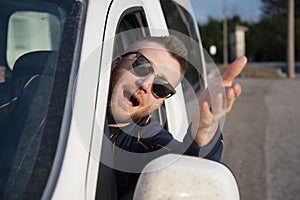 Young man with sunglasses comes out of the car window with his head with his arm raised. Face expression of angry driver arguing