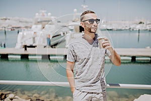 Young man in sunglasses drink coffee to go on summer street with yacht harbor on background. Travel concept