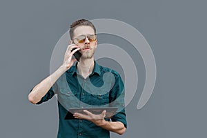 A young man in sunglasses is holding a tablet in his hands and talking on the phone