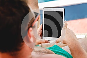 A young man in sunglasses holds a tablet in his hands. Freelancer job concept on the beach and vacation